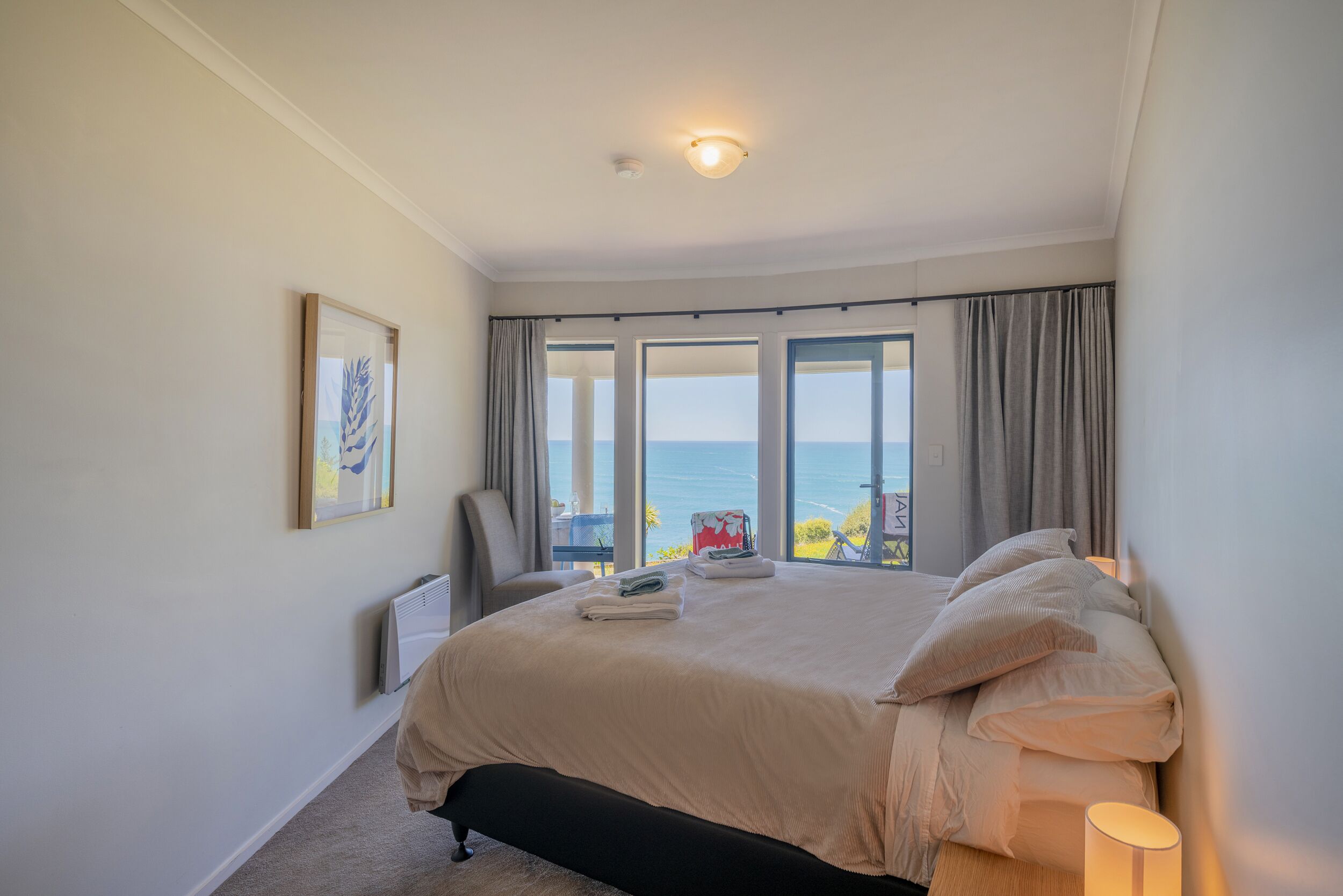 A bedroom with a large bed, a chair, a radiator, and a view of the sea through large windows.