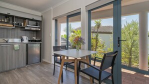 Cozinha moderna com micro-ondas embutido, cafeteira e geladeira. Há uma mesa de jantar com cadeiras e um vaso de flores. O ambiente possui grandes janelas com vista para o verde lá fora.