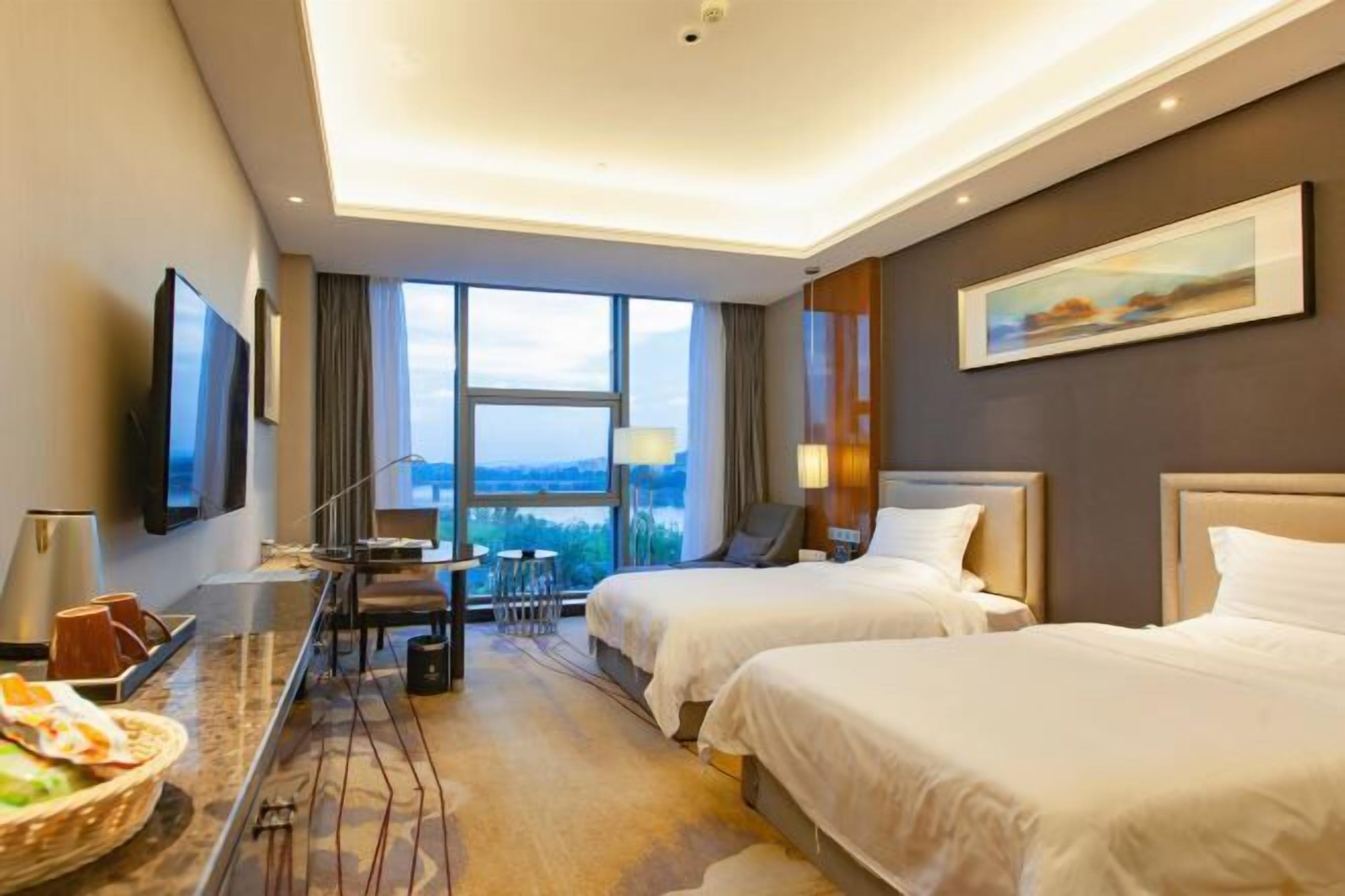 A hotel room with two beds, a desk with a chair, a TV, and a large window offering a view.