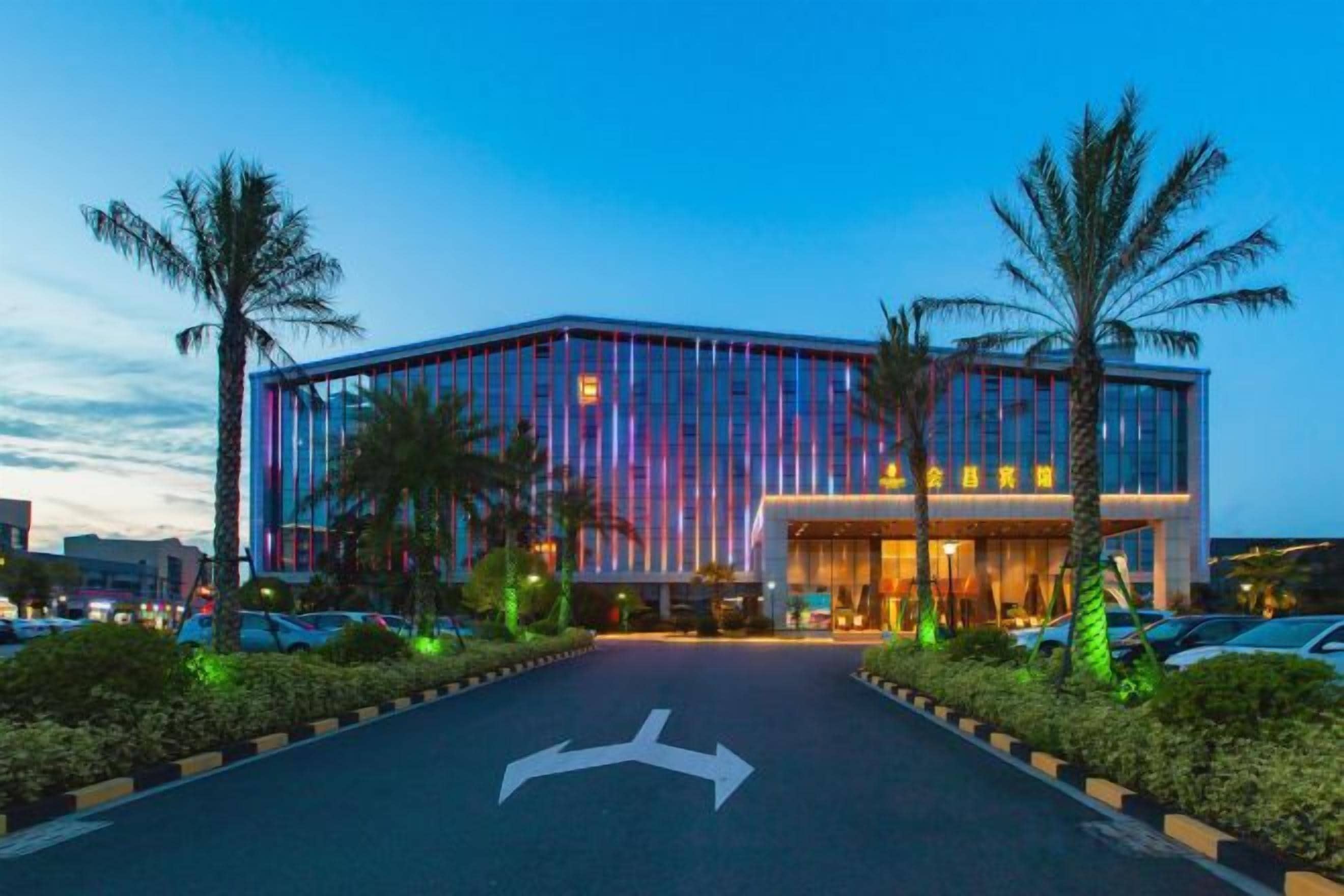 A modern building with a glass facade, a parking lot with directional arrows, and palm trees.