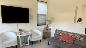 Un salon avec un canapé gris, deux fauteuils blancs, une petite table, un téléviseur à écran plat, une fenêtre avec des stores, une lampe, un panier et une horloge au mur.