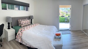 Une chambre à coucher comprenant un lit, des tables de chevet, un bureau et donnant sur un jardin par une porte.
