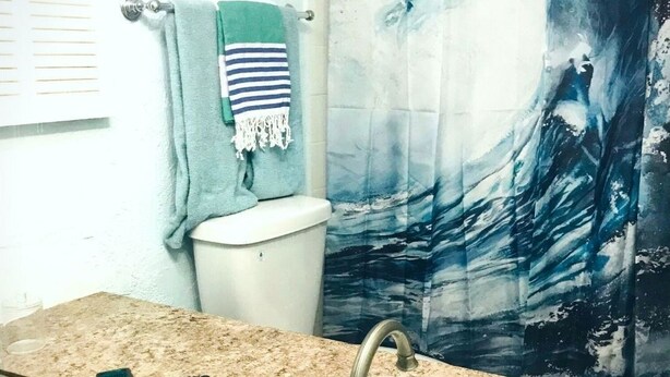 Ein Badezimmer mit einem weißen Waschbecken, zwei Wasserhähnen, einem Spiegel, einem Handtuchhalter mit einem grünen Handtuch und einem Duschvorhang mit Wellenmuster.