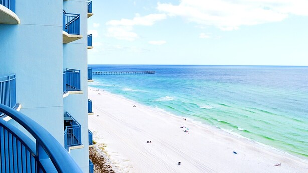 A beach with a pier extending into the ocean, a clear sky, and a building with balconies overlooking the beach. - Sterling Breeze 2Bd/3Ba w/Bunk Room / Near Pier Park /Sleeps 8 (Panama City Beach)