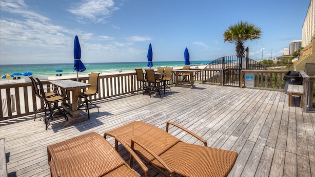 A wooden deck with outdoor seating, a view of the beach, and a palm tree.