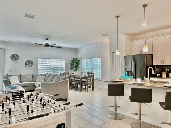 A modern living room with a foosball table, a dining area, and a kitchen with bar stools.