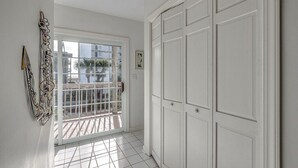 Un pasillo con suelo de baldosas blancas, una puerta blanca con celosía metálica y un armario blanco grande.