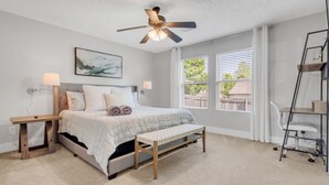 Une chambre à coucher comprenant un lit, des tables de chevet, un banc, un ventilateur de plafond et une fenêtre avec des stores.