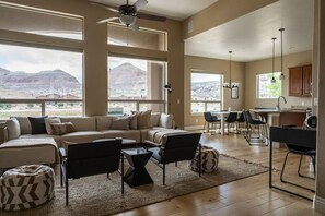 A spacious living room with a large sofa, a coffee table, and a dining area with chairs. There are large windows offering a view of the desert landscape.
