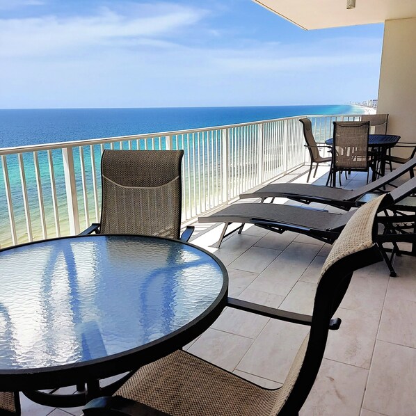 A balcony with a glass table and chairs, overlooking a beach and ocean.