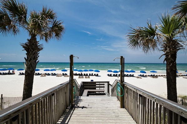 A wooden boardwalk leading to a beach with palm trees and blue umbrellas.