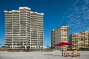 Et højt hotel med flere altaner, beliggende tæt ved en strand med liggestole og parasoller.