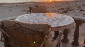 Una mesa redonda con cubierta de mosaico, una silla metálica con cojín estampado y vista al atardecer sobre la playa.