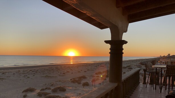 Vista del atardecer desde un balcón con vista a una playa, con una mesa y sillas.