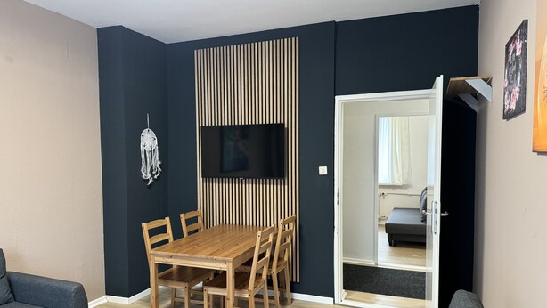Un salon moderne avec une table à manger en bois, deux canapés, un téléviseur fixé à un mur sombre et une porte coulissante donnant sur une autre pièce.