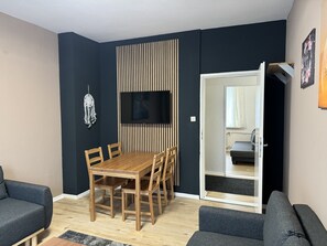 A modern living room with a wooden dining table, two sofas, a TV mounted on a dark wall, and a sliding door leading to another room.