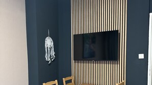 Un coin repas avec une table et des chaises en bois, un téléviseur fixé au mur, lequel est recouvert de lambris en bois verticaux, et un plafond blanc.