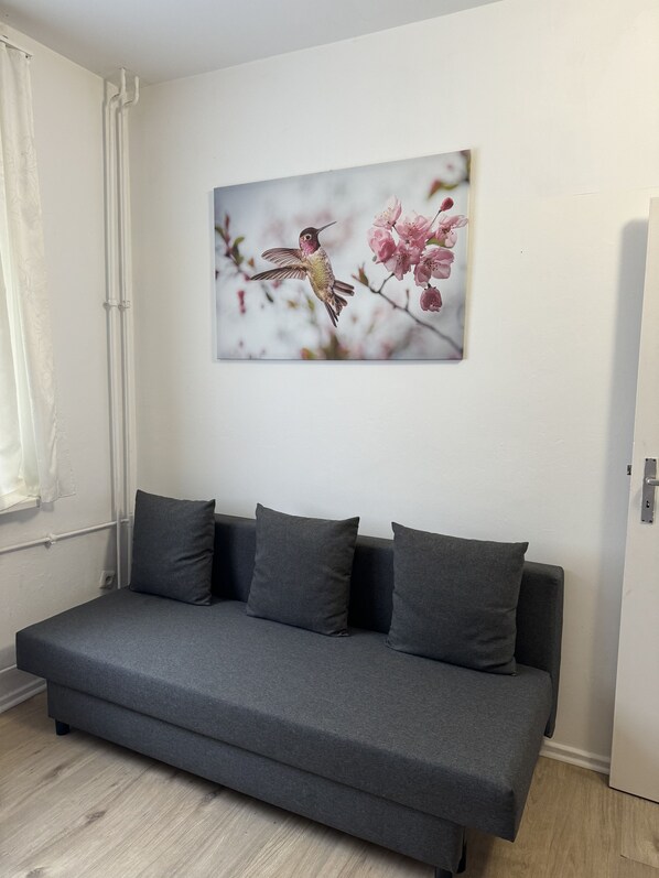 A grey sofa with three cushions, a wall-mounted picture of a hummingbird, and a window with white curtains.