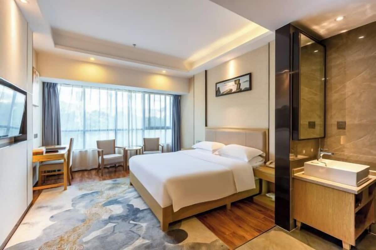 Ein modernes Hotelzimmer mit einem großen Bett, einem Schreibtisch, einem Stuhl, einem Badezimmer mit Waschtisch und einem großen Fenster mit Vorhängen.