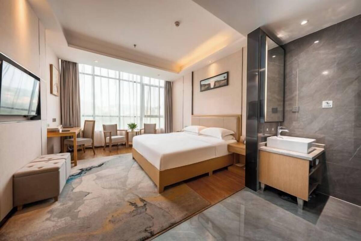 Ein modernes Hotelzimmer mit einem großen Bett, einem Schreibtisch, einem Sessel, einem kleinen Tisch, einer Couch, einem Badezimmer mit Waschbecken und Spiegel sowie einem großen Fenster.