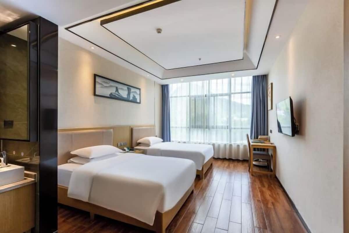 Ein Hotelzimmer mit zwei Betten, einem großen Fenster mit Vorhängen, einem Flachbildfernseher und einem Holzboden.