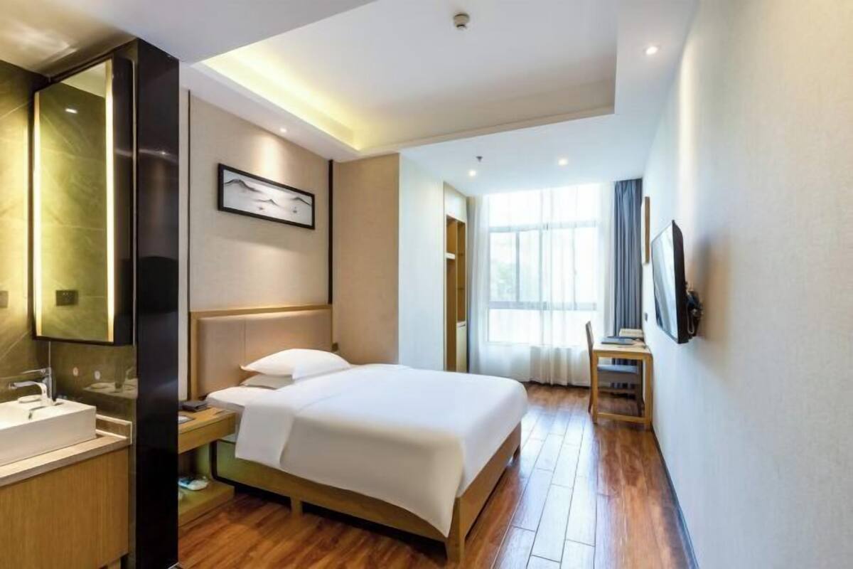 Ein Hotelzimmer mit Bett, Nachttisch, Spiegel, Wandfernseher und einem fenster mit Vorhängen.