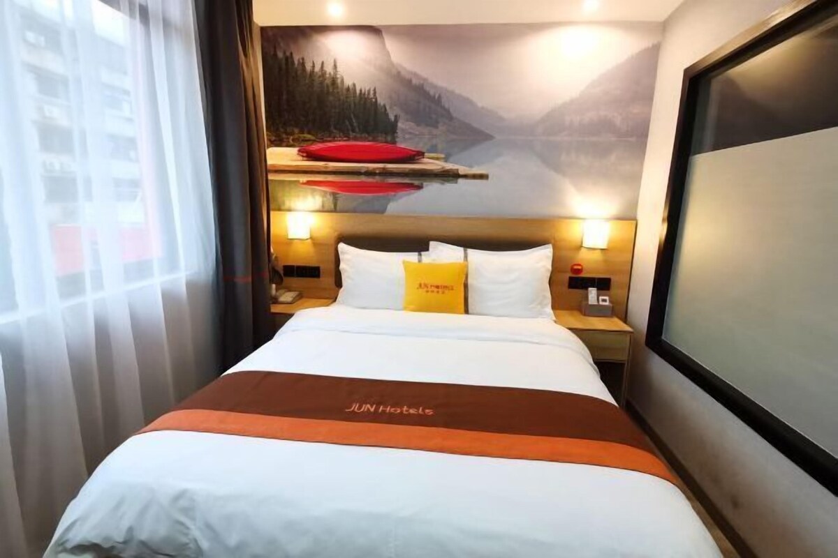 Habitación de hotel con cama, mesita de noche, televisor montado en la pared y ventana con cortinas.