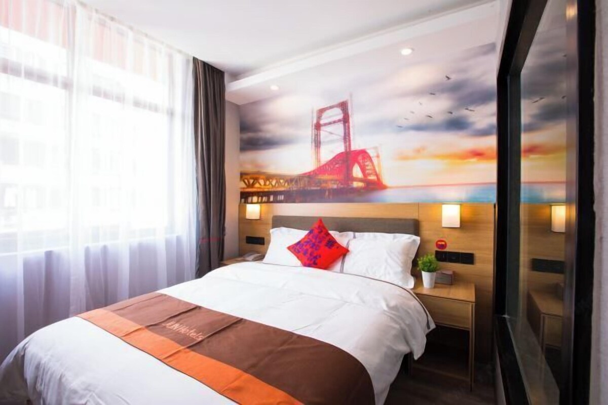 Una habitación de hotel con cama, mesita de noche, lámpara de pared y ventana con cortinas.