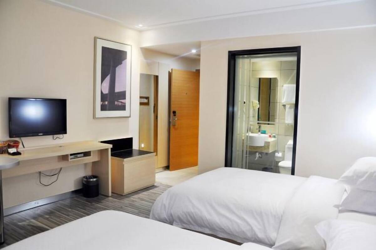 A hotel room with a bed, a TV, a desk, a mirror, and a bathroom visible through a glass door.