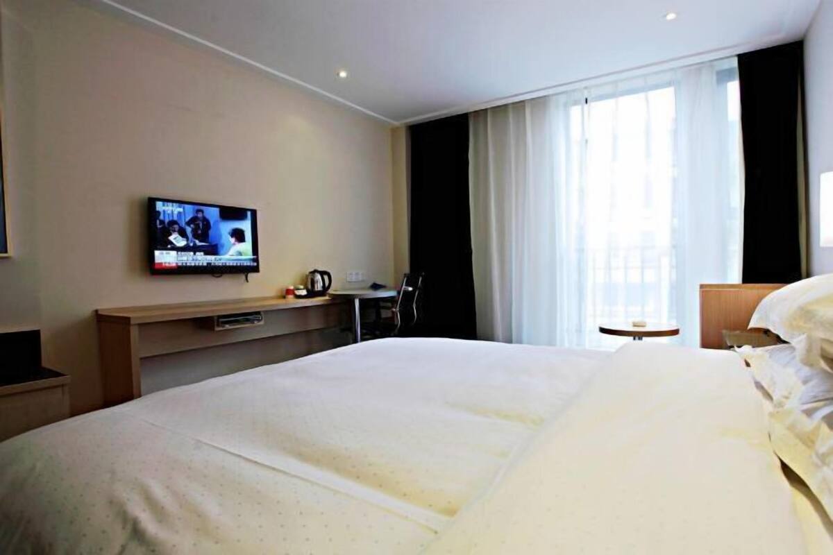 A hotel room with a large bed, a television on the wall, a desk with a chair, and a window with curtains.