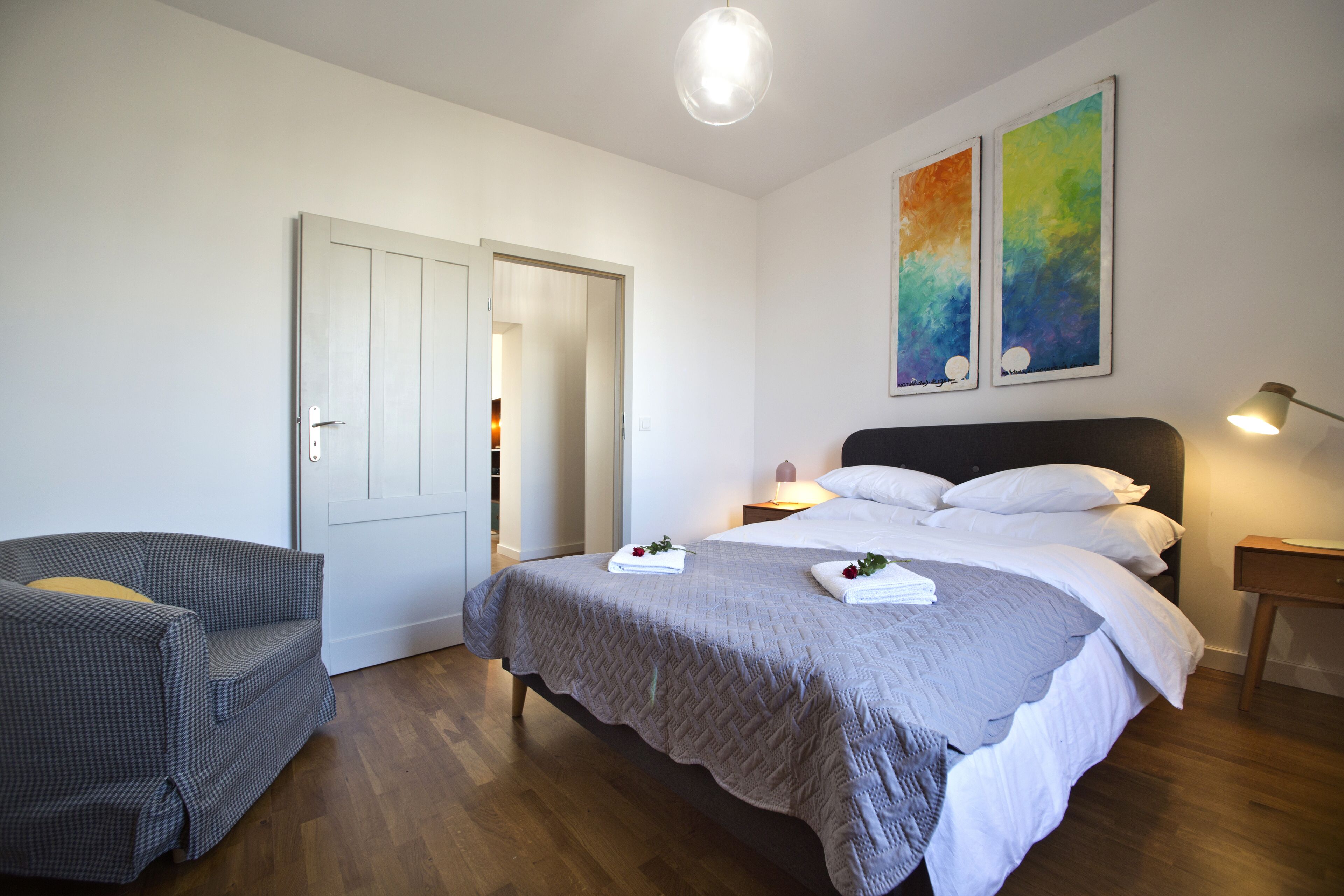 Een slaapkamer met een bed, een stoel, een nachtkastje met een lamp en twee schilderijen aan de muur.