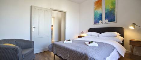 Een slaapkamer met een bed, een stoel, een nachtkastje met een lamp en twee schilderijen aan de muur.