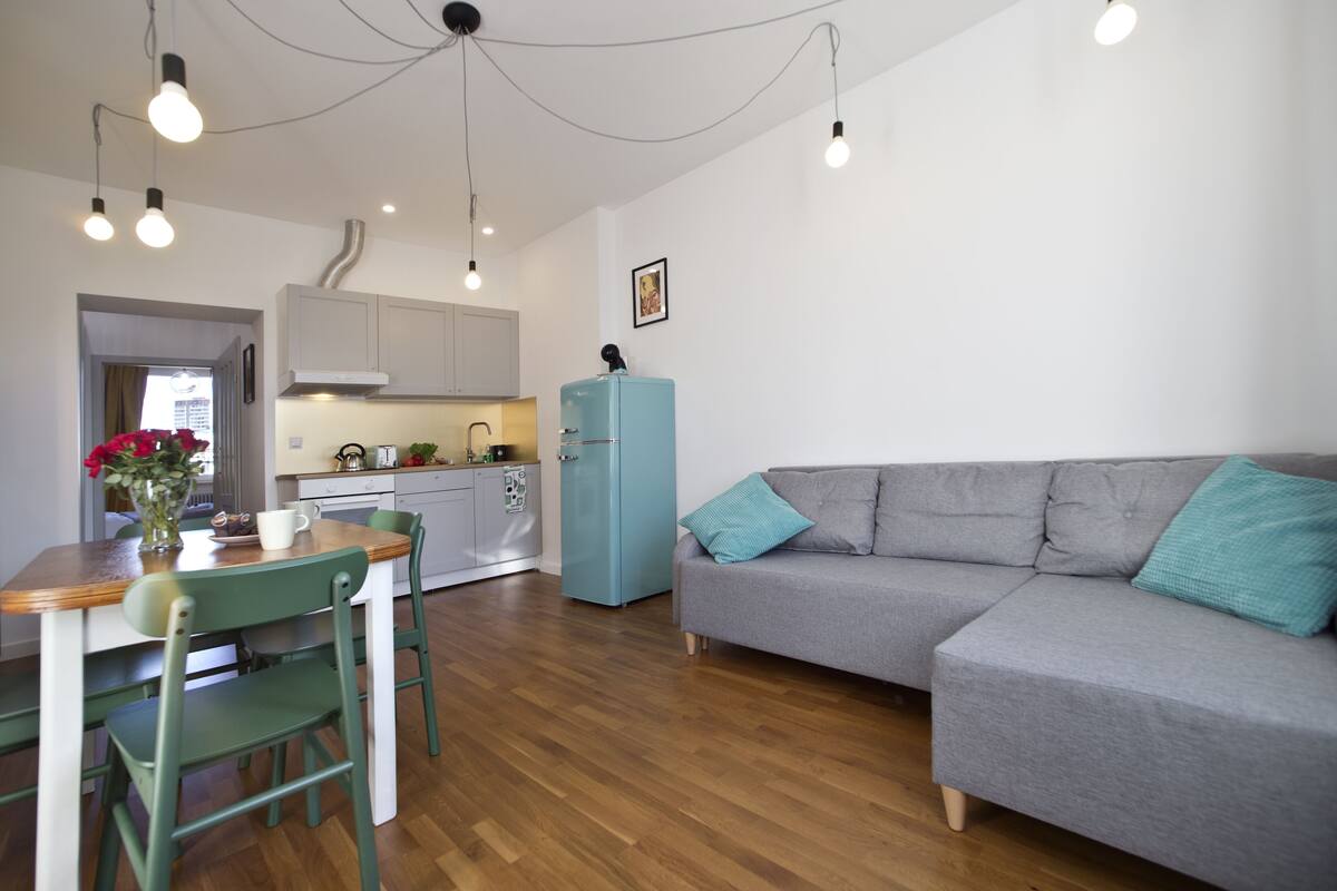 Una sala moderna con un sofá gris, una mesa de comedor de madera con sillas, un refrigerador azul claro e iluminación colgante.