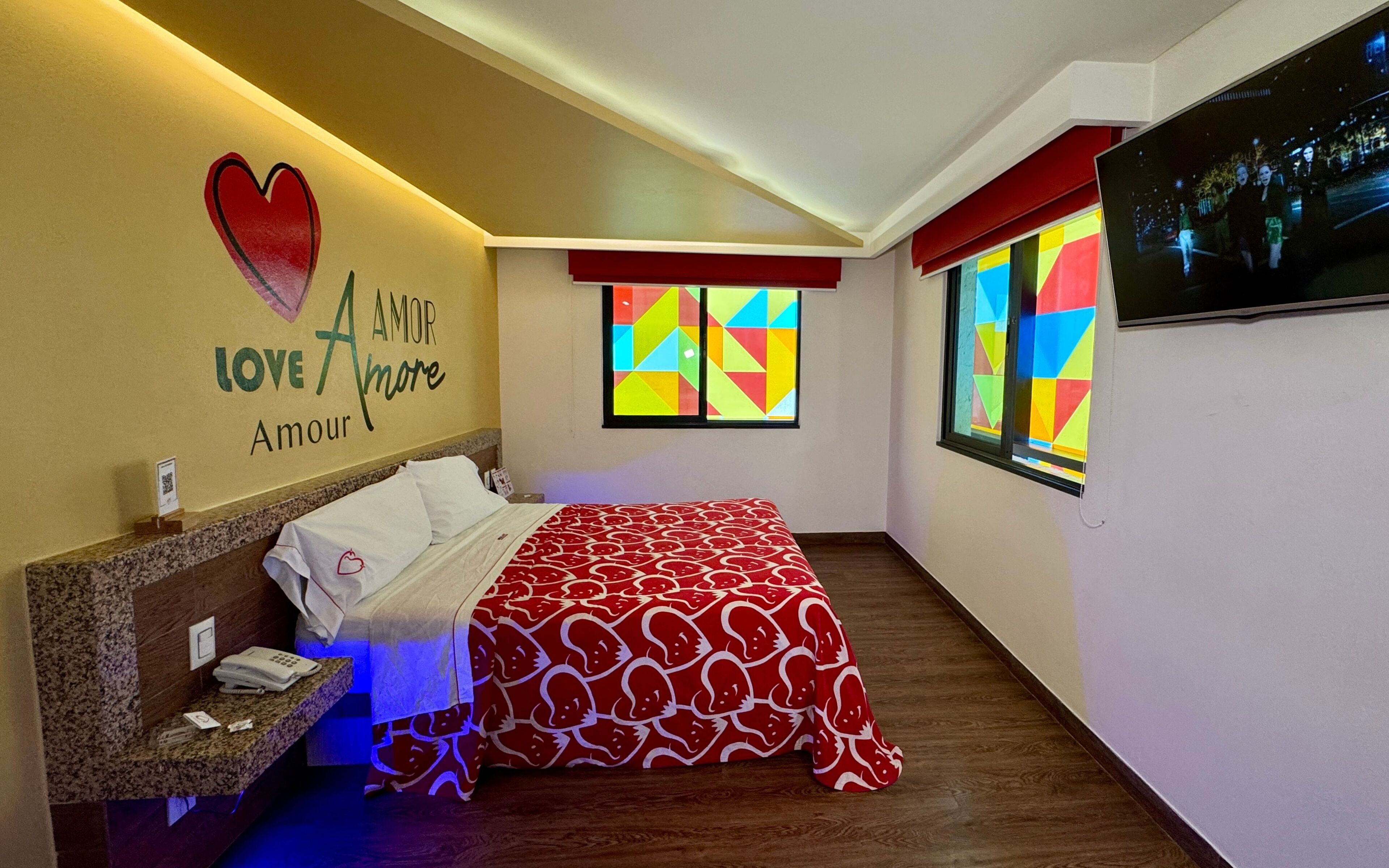 Habitación de hotel con una cama, una pared con un diseño en forma de corazón y las palabras "Love Amore", una ventana con un motivo geométrico colorido, y un televisor en la pared.