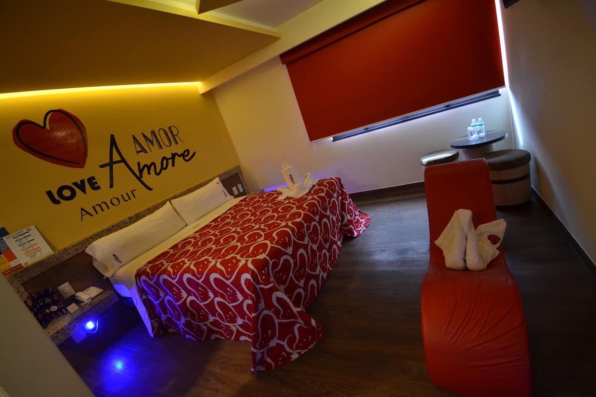 Hotelkamer met een bed, een rode stoel, een tafeltje met waterflesjes, en een muur met een hart en de tekst 'Love Amore'.