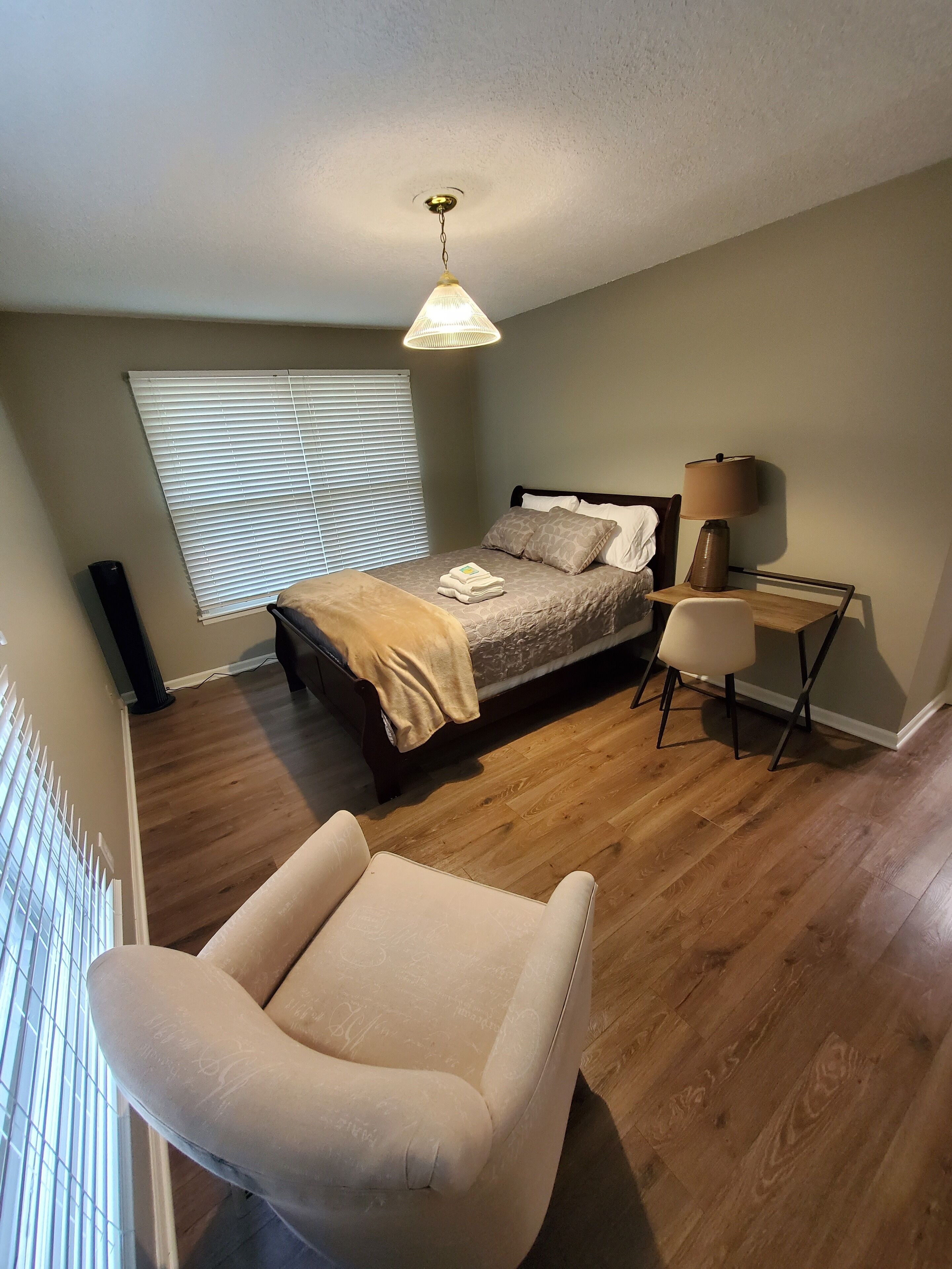 A bedroom with a bed, bedside lamp, chair, and a window with blinds.