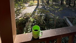 Une terrasse en bois avec une tasse verte, donnant sur une zone boisée, sous un ciel dégagé où la lumière du soleil filtre à travers les arbres.