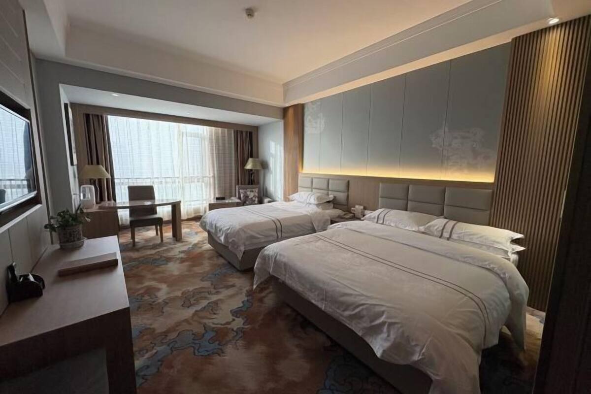 Habitación de hotel con dos camas, un escritorio, una silla, una lámpara y un ventanal grande.