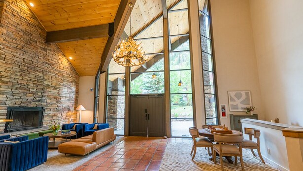 A spacious living room with a stone fireplace, large windows, and a high ceiling with exposed beams.