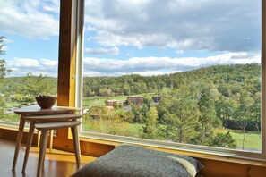 A room with a large window offering a view of a forested area and buildings. There is a wooden table with a bowl on it. - Cliff’s End • Ski & Golf Views • Fire Pit • Game Room (Basye)