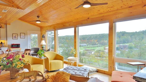 Una habitación amplia con grandes ventanales, techo de madera y muebles modernos, incluyendo un sillón amarillo, un sofá y una mesa de comedor con una escultura de madera decorativa.
