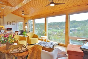 En rummelig stue med store vinduer, træloft og moderne møbler, herunder en gul lænestol, en sofa og et spisebord med en dekorativ træskulptur.
