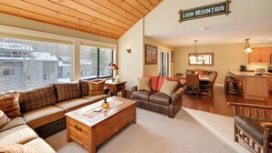 A spacious living area with a wooden ceiling, a large window, a sofa, a coffee table, and a kitchen area with a sign reading "LOON MOUNTAIN". - Lincoln Condo w/ Balcony & View < 2 Mi to Loon Mtn (Lincoln)