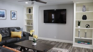 A modern living room with a black leather sofa, a coffee table, a TV mounted on the wall, and a white shelving unit with decorative items. - Chisholm Creek by Top Golf 2 (Oklahoma City)