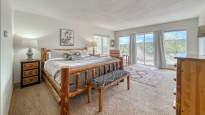 A bedroom with a wooden bed frame, a bench, a nightstand, a chair, and a view of the outdoors through large windows. - Beautiful Home with Breathtaking Views! (Nathrop)