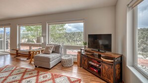 A spacious living room with a sofa, ottoman, and a TV on a wooden stand. Large windows offer a view of the outdoors. - Beautiful Home with Breathtaking Views! (Nathrop)