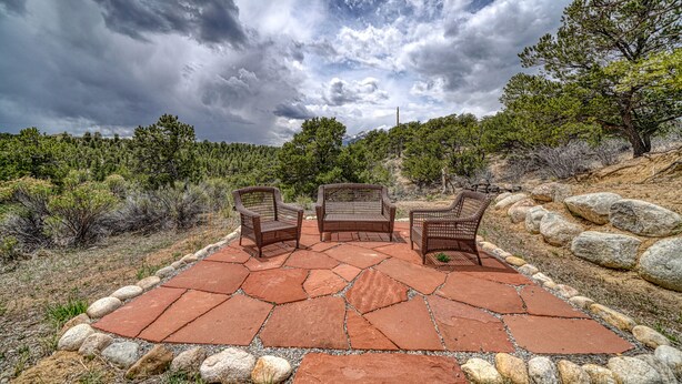 A patio with three wicker chairs and a table, surrounded by a stone pathway and large rocks, set against a backdrop of trees and a cloudy sky. - Beautiful Home with Breathtaking Views! (Nathrop)