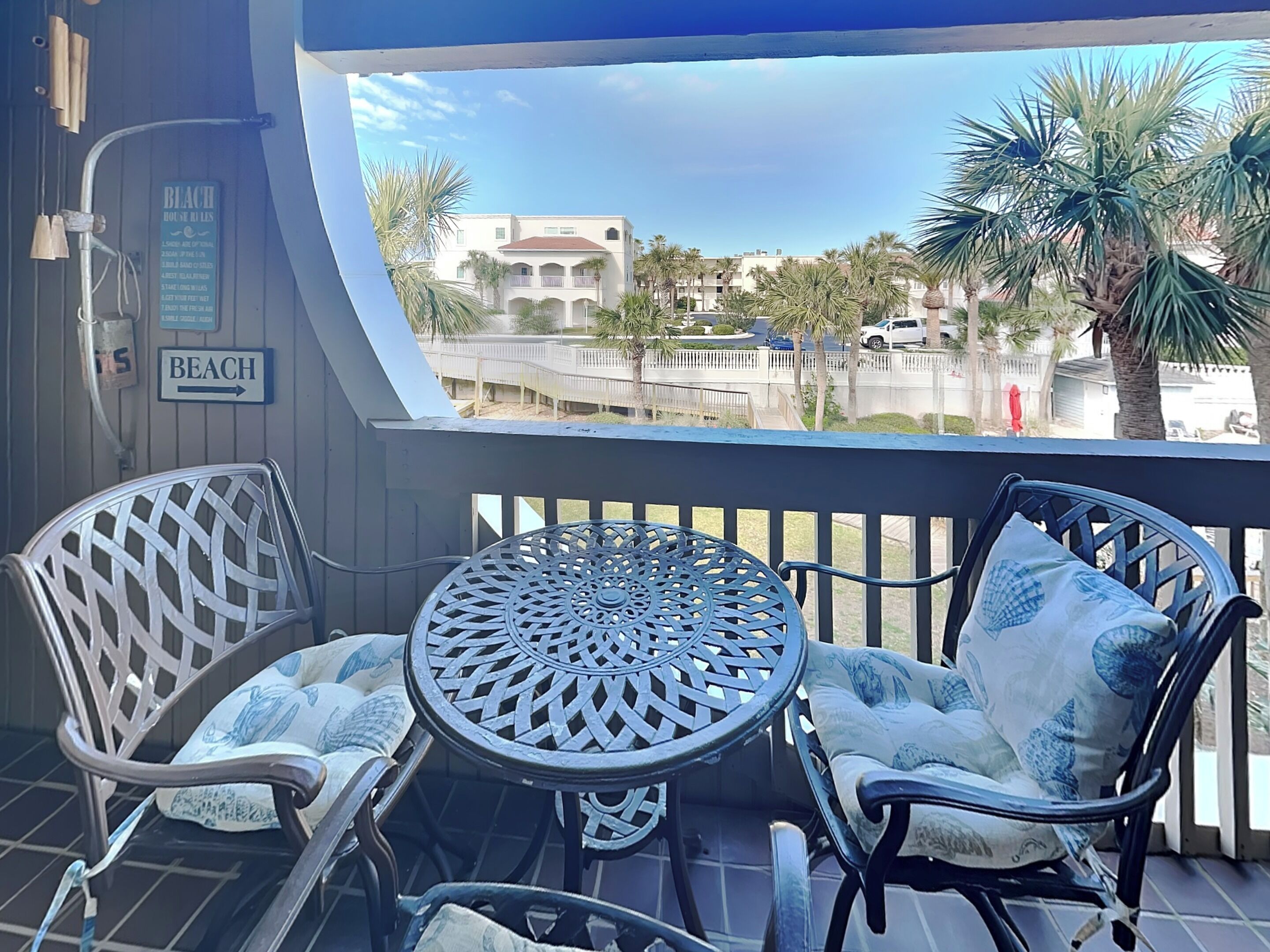 A balcony with a view of a beach and palm trees. There are two wicker chairs with cushions and a metal table. A sign on the wall reads "BEACH."