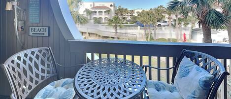 A balcony with a view of a beach and palm trees. There are two wicker chairs with cushions and a metal table. A sign on the wall reads "BEACH."