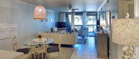 A spacious living area with a dining table, chairs, a sofa, and a TV. There is a balcony with a view of palm trees.